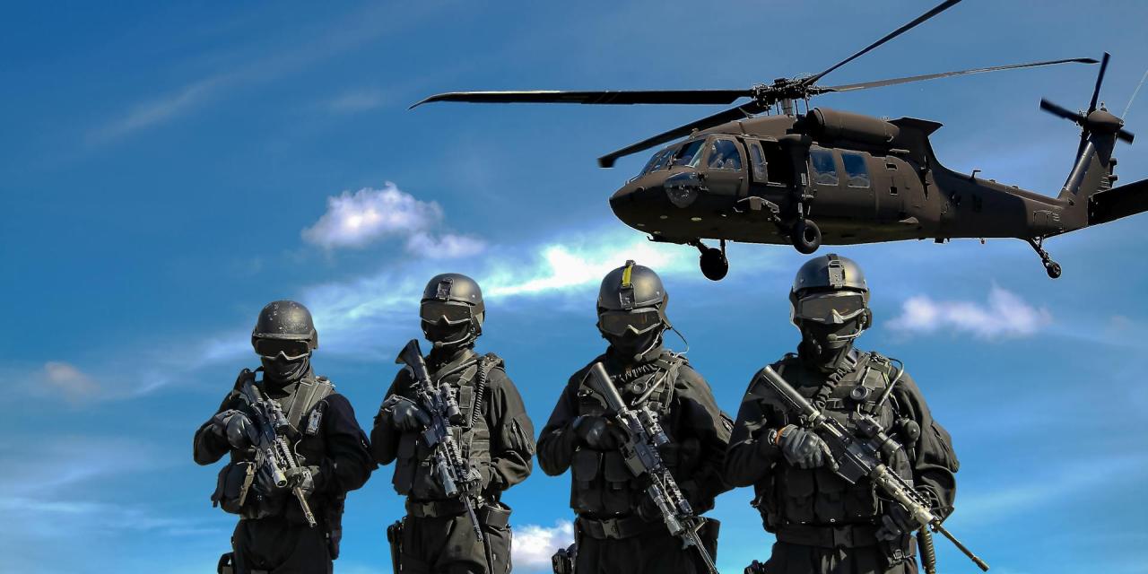 Vier Personen mit Schutzausrüstung und Gewehren. Im Hintergrund ein Hubschrauber vor blauem Himmel.