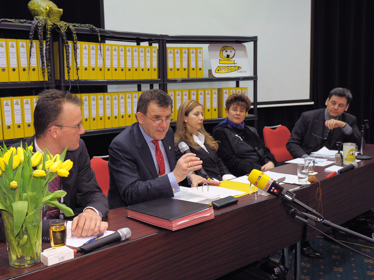 Rena Tangens and padeluun at a table at the press conference in front of a shelf with a lot of yellow folders