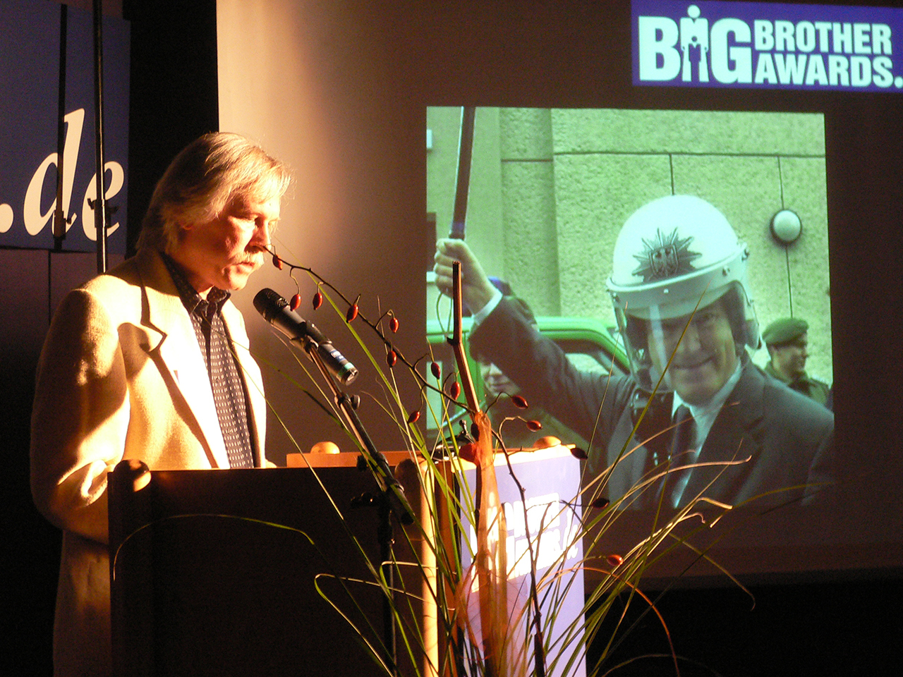 Rolf Gössner at the lectern at the BigBrotherAwards. On the canvas Otto Schily in police uniform