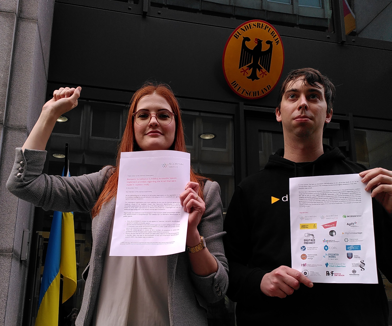 Ella von EDRi und Konstantin von Digitalcourage halten den offenen Brief an die deutsche Bundesregierung hoch und stehen dabei vor der Ständigen Vertretung Deutschlands an die EU.
