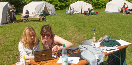 Zwei Personen sitzen an einem Tisch vor einem Laptop. Im Hintergrund stehen Zelte.