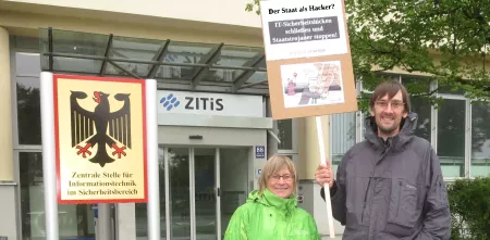 Zwei Personen mit einem Demoschild vor der Zentralen Stelle für Informationstechnik im Sicherheitsbereich. 