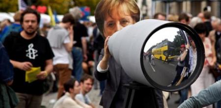 Ein riesiges Fernglas auf einer Demo aufgebaut. Dahinter steht eine Person mit einer Angela-Merkel-Maske.