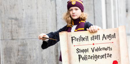 Ein Kind, das verkleidet ist wie eine Figur aus Harry Potter. In der Hand ein Schild und ein Zauberstab. Auf dem Schild steht: „Freiheit statt Angst. Stoppt Voldemorts Polizeigesetze“.