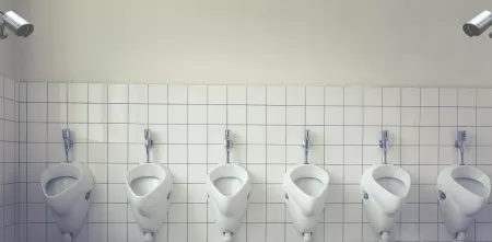 Mehrere Pissoirs nebeneinander. In die Ecken oben rechts und links wurden zwei Überwachungskameras per Fotomontage eingefügt.