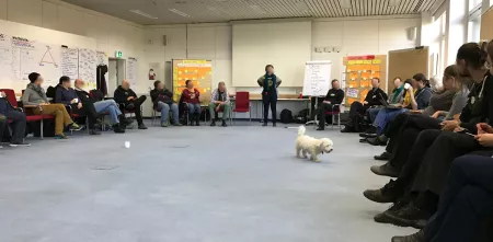 Ein Sitzkreis mit vielen Menschen, eine Person steht an einer Flipchart. 