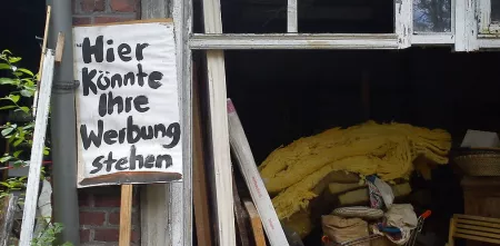 Eine vollgestellte Garage. Davor ein Schild mit dem Schriftzug: „Hier könnte ihre Werbung stehen“.