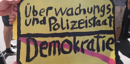 Ein Demoschild, das aussieht wie ein Ortsausgangschild (schwarze Schrift auf gelben Grund). Oben: Überwachungs- und Polizeitstaat. Unten durchgestrichen in rot: Demokratie.
