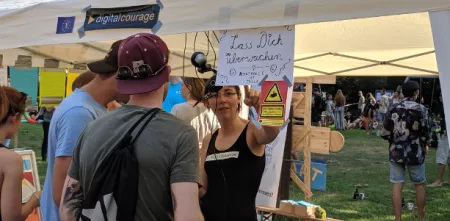 Zwei Personen steht vor einem Zelt-Pavillion. Vor ihnen ein Schild mit der Aufschrift „Lass dich überraschen!“
