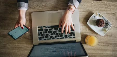 Draufsicht eines Laptops mit einer Person, die tippt und gleichzeitig ein Smartphone in der Hand hält.