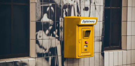 Montage: Ein gelber Kasten an einer Hauswand. Darauf steht „Digitalzwang“.