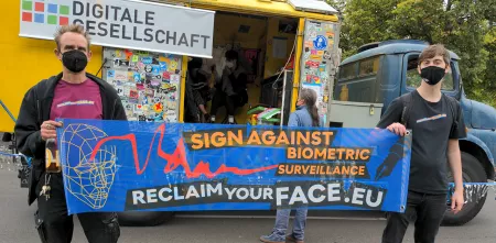 Zwei Menschen halten ein Reclaim Your Face Banner hoch und stehen vor einem Lautsprecherwagen bei der unteilbar-Demo in Berlin 2021.