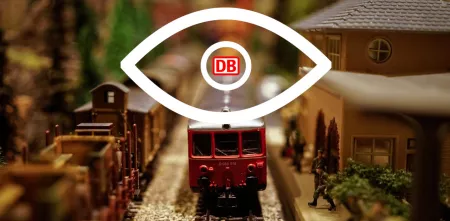 Ein großes Auge, dessen Pupille aus dem Logo der Deutschen Bahn AG besteht, überblickt einen Miniaturbahnhof.