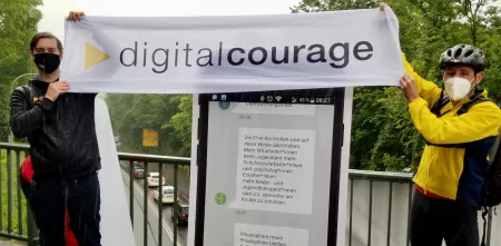 Zwei Personen halten ein Banner mit dem Logo und Schriftzug von Digitalcourage vor einem menschenhohen Smartphone-Aufsteller