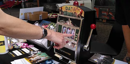 Ein Arm zeigt auf einen selbstgebauten einarmigen Banditen. Drumherum liegt Infomaterial von Digitalcourage.