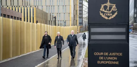 Drei Personen laufen auf die Kamera zu. Rechts im bild ist ein Schild, welches den Europäischen Gerichtshof ausweist.