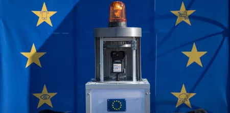 Vor einer EU-Flagge mit Sternen, die Überwachungssymbole in sich haben, steht ein Apparat. Der Apparat hat mittig eine Handyhalterung, oben eine Alarmleuchte und unten ein Gehäuse aus Plexiglas mit Metallprofilen.