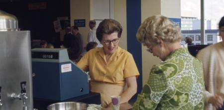 Kasse in einer Cafeteria der 1970er Jahre. Zwei ältere Frauen im Bezahlvorgang.