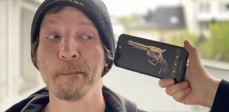 Ein Mann mit bart und Mütze bekommt ein Smartphone an die Schläfe gehalten auf dem ein altertümlicher Revolver abgebildet ist.