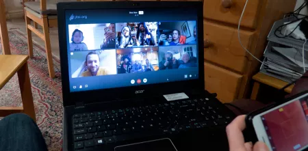 Laptop auf dem Schoß, darauf läuft eine Jitsi-Konferenz, eine Hand mit Smartphone ragt leicht ins Bild.