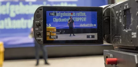 Ein Mensch ist auf dem Bildschirm einer Videokamera zu sehen. Er steht, mit einem Megaphon bewaffnet, vor einem Protestbanner.