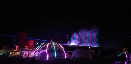 Bunte Farben erstrahlen an und über Zelten in der Dunkelheit. Entlang einer Anhöhe ergeben Richtung Himmel gerichtete Strahler ein blau/lila gestreiftes Muster.