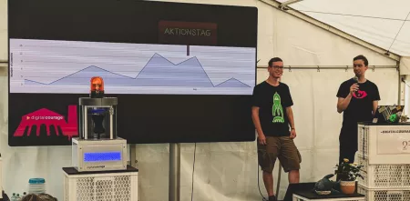 Zwei Menschen stehen auf einer Bühne. Links im Vordergrund ist ein Apparat (der „Chatkontrolle-Prototyp“) und im Hintergrund ist ein Kurvendiagram in einer Präsentation abgebildet. Bei den zwei kurz aufeinander folgenden Höhepunkten der Kurve ist eine Markierung mit „Aktionstag“.