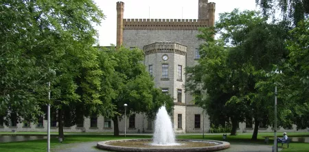 Die Ravensberger Spinnerei in der Frontalansicht. Links und rechts große Bäume. Im Fokus ein Springbrunnen direkt vor dem historischen Gebäude.