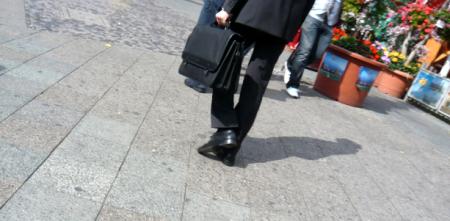 Eine Geschäftsperson im Anzug mit schwarzem Aktemkoffer auf einer Straße. Nur der Beinbereich ist zu sehen.