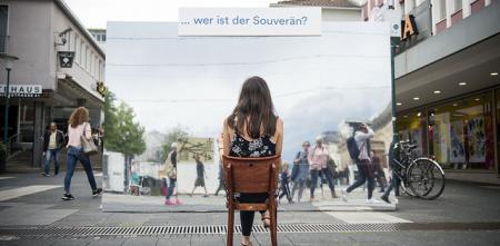 Eine Person die auf einem Stuhl in einer Innenstadt vor einem Plakat sitzt. Darauf die Frage: „Wer ist der Souverän?“