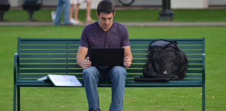 Eine Person sitzt auf einer Parkbank. Auf dem Schoß steht ein Laptop, auf der Bank liegt ein aufgeschlagenes Buch.