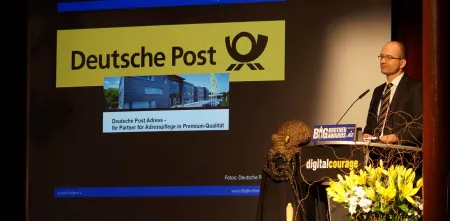 Sönke Hilbrans am Redner.innenpult der BBAs 2013. Im Hintergrund das Logo der Deutschen Post.