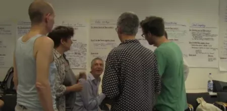 Mehrere Personen stehen im Kreis. Im Hintergrund viele Flipchart-Notizen.