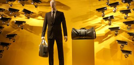 Eine Schaufensterpuppe in Anzug mit zwei Taschen vor gelben Hintergrund, auf die sehr viele Überwachungskameras gerichtet sind.