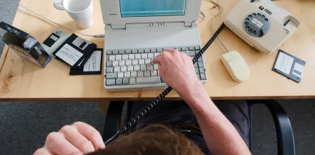 Draufsicht wie eine Person auf einem sehr alten Laptop tippt. Daneben Disketten und ein Wählscheibentelefon.