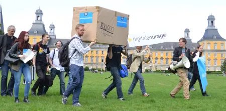 Mehrere Personen tragen ein riesiges Paket über eine Wiese. Auf der Box steht 500.000 Eingaben.