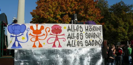 Ein Banner mit 3 Affen. Daneben der Text: „Alles hören! Alles sehen! Alles sagen?“