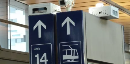 Detailaufnahmer von Hinweisschildern am Bahnhof. Darüber eine Überwachungskamera.