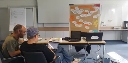 Zwei Personen sitzen auf Stühlen und schauen auf einen Laptop-Bildschirm. Rechts daneben steht eine Flipchart.