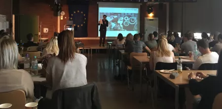 Schüler.innen an Tischen in einer Klasse. Auf einem Podium Friedemann Ebelt. Das Beamer-Bild zeigt das Logo von Digitalcourage und eine Grafik eines Kopfes bestehend aus Zahnrädern.