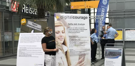Leena Simon mit einer Maske neben einem Plakat „Selfie statt Analyse“. Im Hintergrund sind mehrere Polizist.innen. Aufnahmeort: Ein Bahnhof.