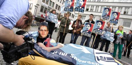 Aktion in der Innenstadt: Eine Person mit Edward-Snowden-Maske liegt in einem Bett. Im Hintergrund Menschen mit Edward-Snowden-Schildern.