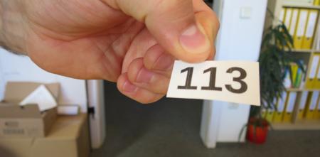 Detailaufnahmer einer Hand, die einen Zettel mit einer Zahl (113) in der Hand hält.