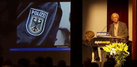 Rolf Gössner am Redepult während einer BBA-Laudatio. Beamerbild: Wappen der Polizei.