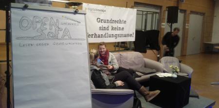 Ein Flipchart mit dem Text "Open Sofa – Lesen gegen Überwachung". Im Hintergrund zwei Personen auf einem Sofa sitzend.