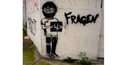 Ein Graffiti einer Person mit einer Art Fotoapparat in den Händen. Daneben steht das Wort „Fragen“.