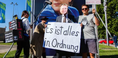 Mehrere Aktivist.innen vor einem Plakat mit Angela Merkel. Sie halten Demo-Schilder („Es ist alles in Ordnung“) und ein Mikrofon in der Hand.
