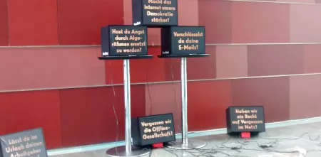 Installation: Mehrere Fernseher vor roter Wand. Darauf sind diverse Texte abgebildet.