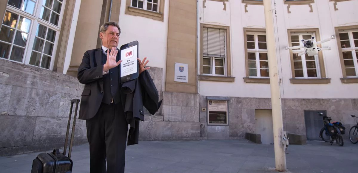 padeluun vor dem Landgericht Frankfurt/Main, der von einer Drohne verfolgt wird. Auf einer Smartphone-Attrappe steht: „DB Schnüffel-Navigator. Thank you for tracking with Deutsche Bahn“.