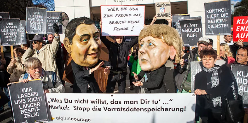 Aktivist.innen mit diversen Bannern. Im Hintergrund Pappmaché-Köpfe von Angela Merkel und Sigmar Gabriel.